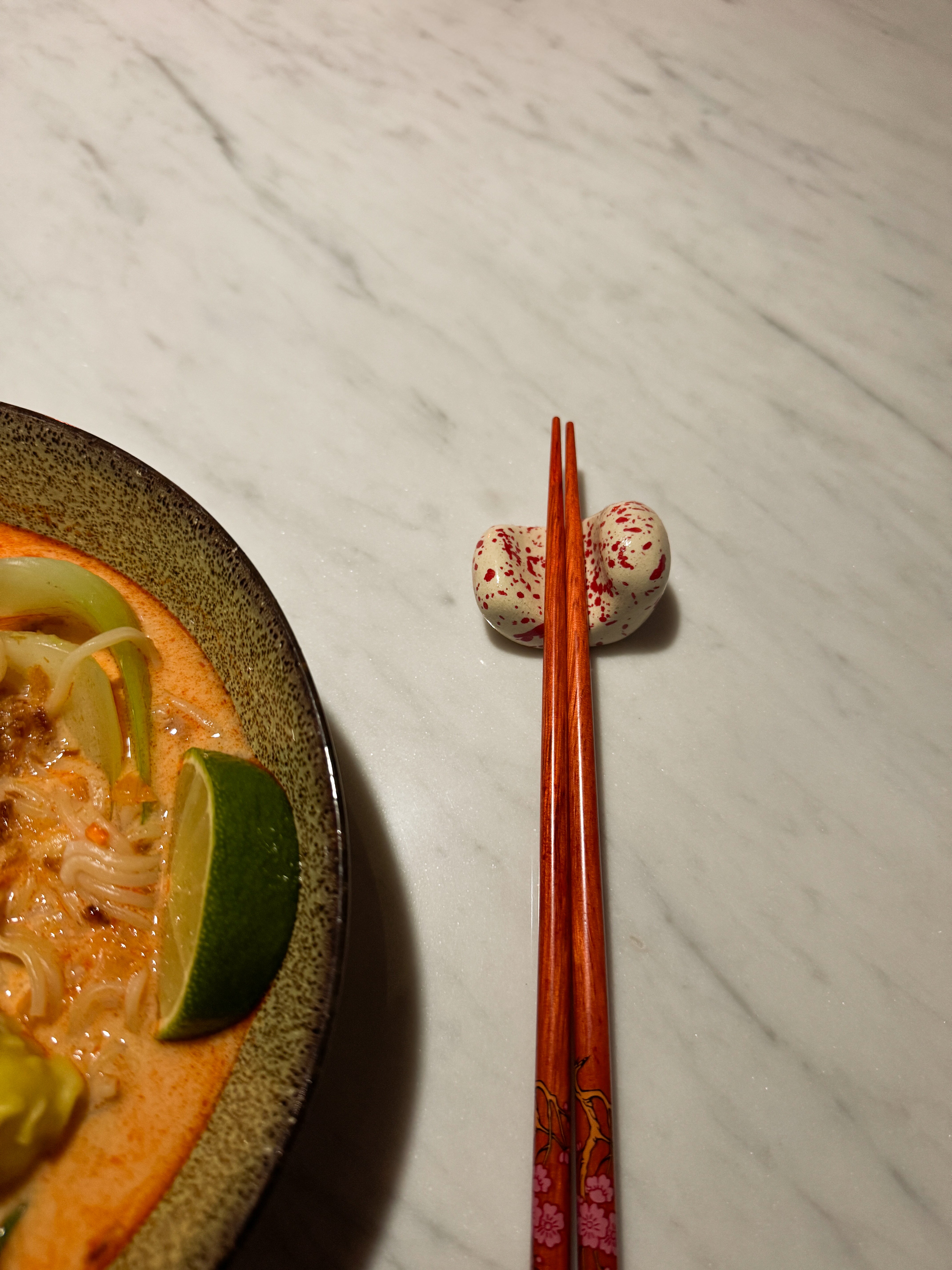 Handgemaakt keramiek eetstokjeshouder in het beige met rode spetter afgebeeld met rode eetstokjes erop en een bord noodlesoup ernaast op een marmeren tafel