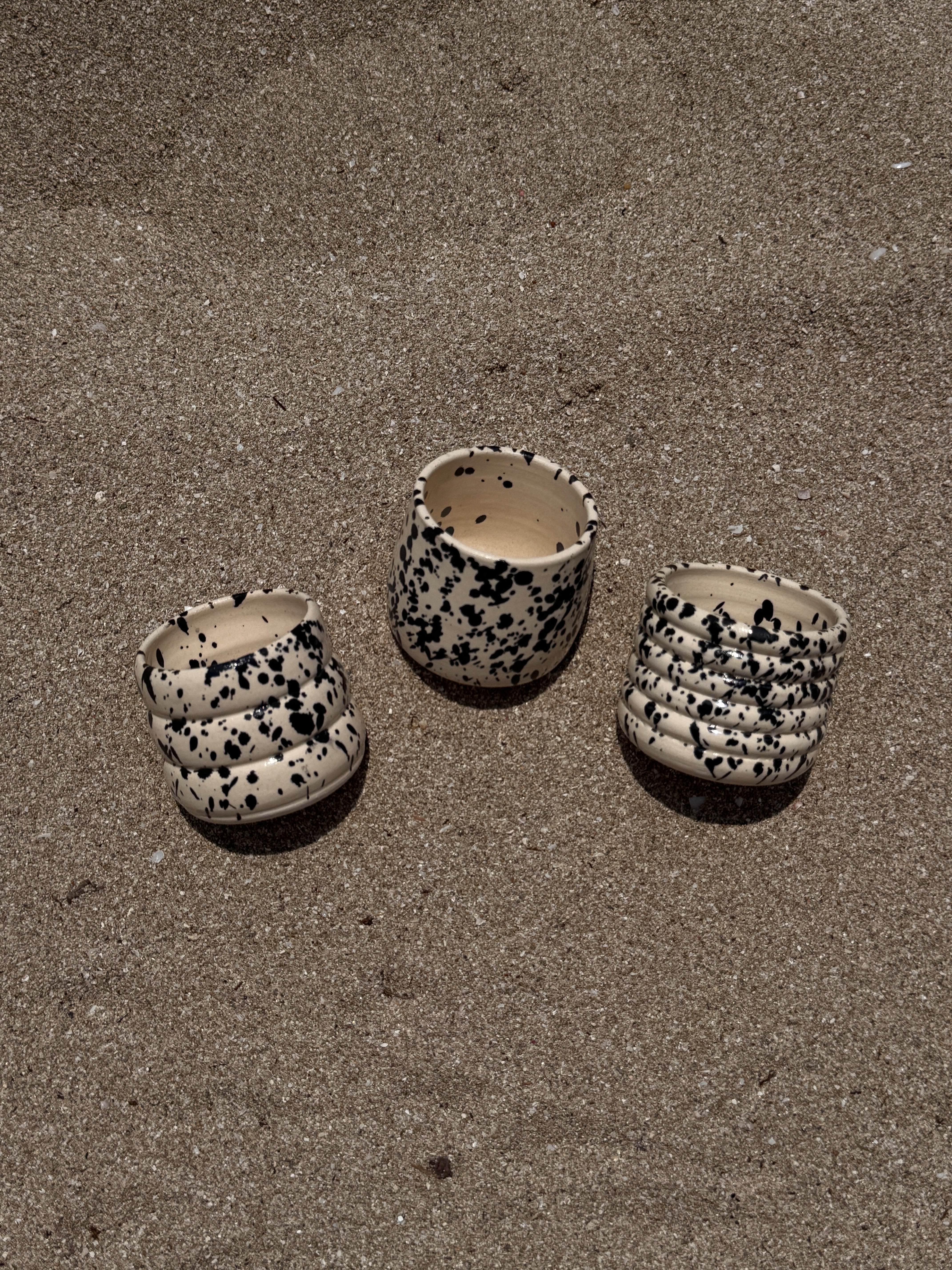 drie handgemaakte keramiek mokjes in drie verschillende structuren allen met beige kleur en zwarte splatters liggend in het zand 